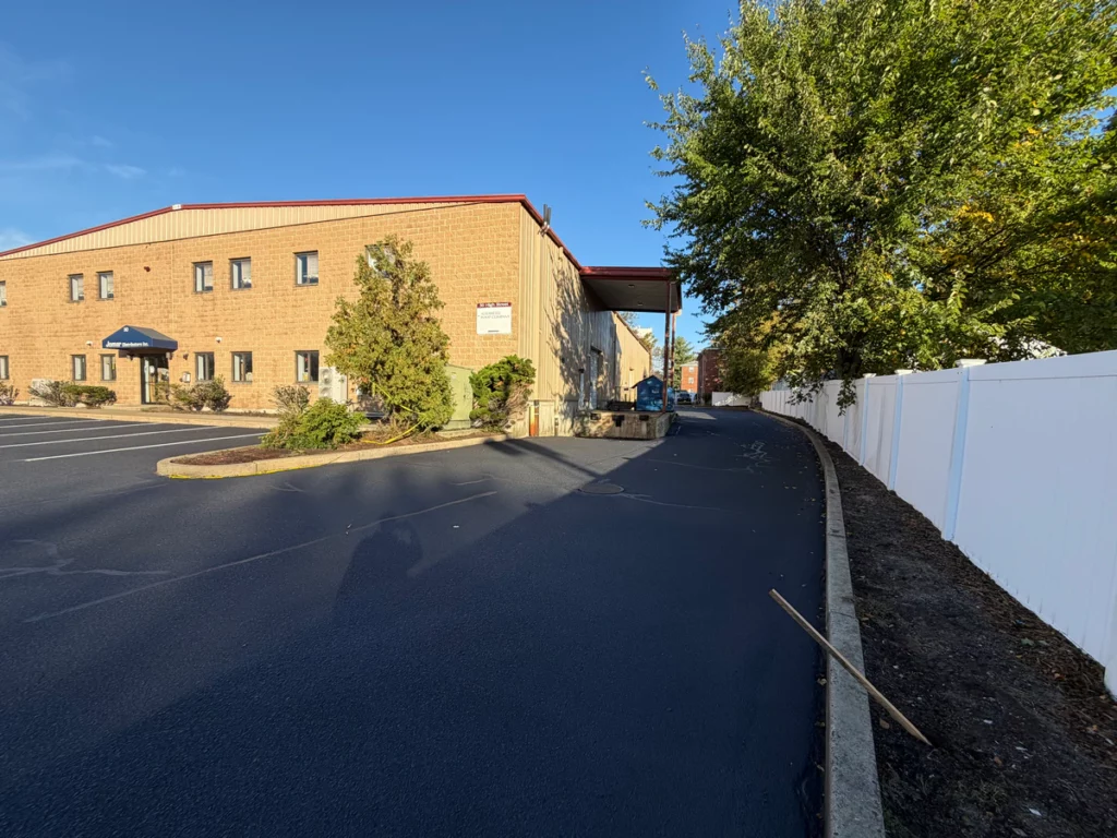 Freshly sealcoated service lane in Woburn, MA curving along a white vinyl fence to a covered loading dock.